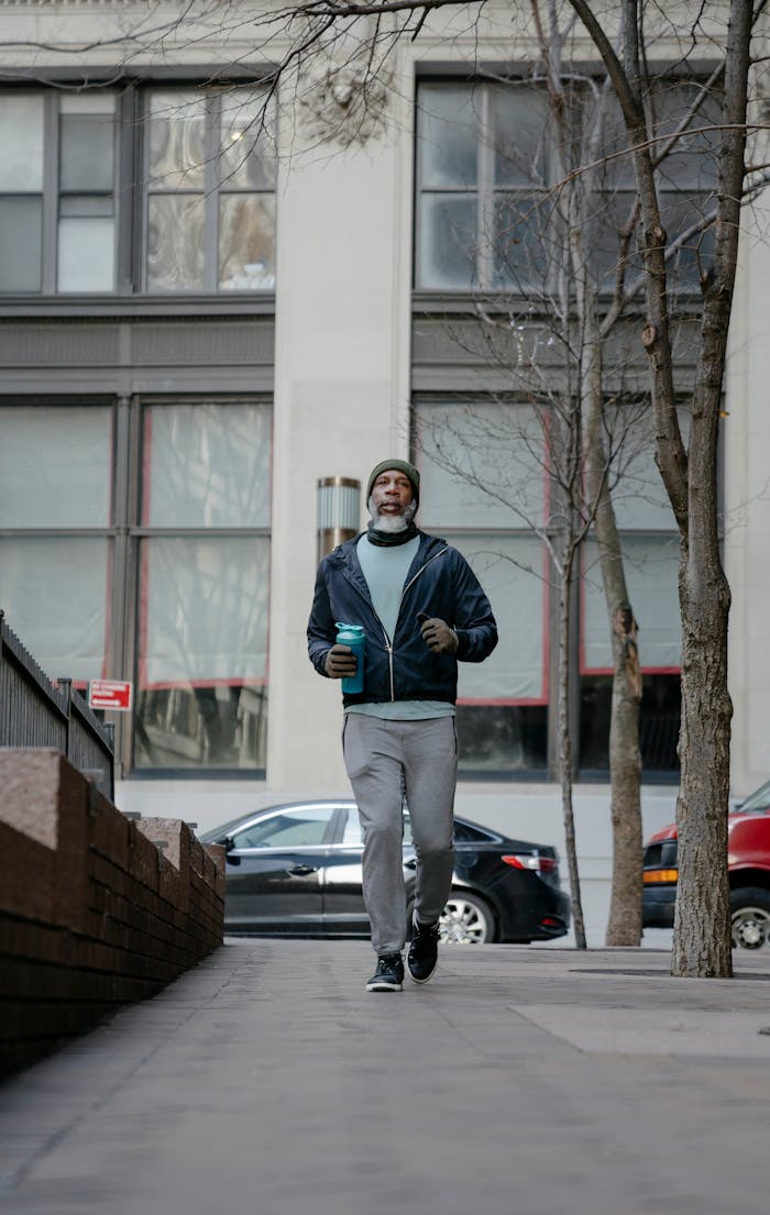 Full body of senior African American male wearing sportswear and with bottle in hand jogging on paved pathway in city